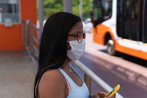 Jonge vrouw die gezichtsmasker draagt die telefoon in busstation gebruikt.
