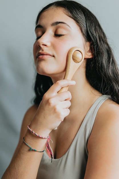 Jonge vrouw die gezichtsmassage toepast met houten rol Druk uitoefenen om de bloedsomloop te verbeteren Nieuwe methoden voor huidverzorging en dagelijkse rituelen Ontspannen zelfzorgdag Portret in studio met grijze achtergrond