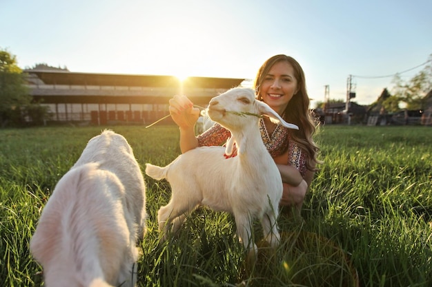 Jonge vrouw die gras voedt met geitenkinderen, groothoekfoto met sterke achtergrondverlichting en zon over boerderij op de achtergrond