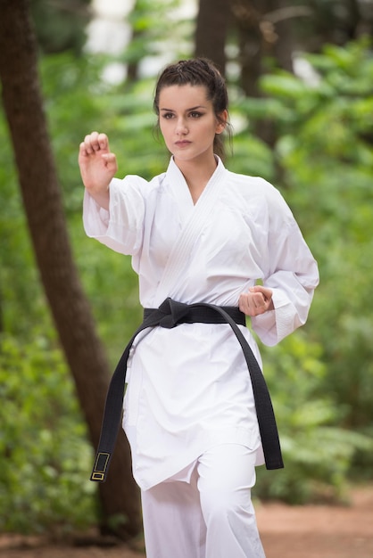 Jonge vrouw die haar karatebewegingen beoefent in bebost bosgebied Witte kimono zwarte band