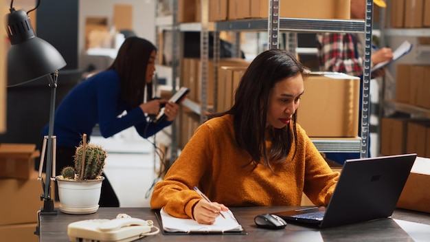 Jonge vrouw die handelswaar in stapels organiseert, retailproducten in pakketten stopt voor distributie. Vrouwelijke ondernemer die laptop gebruikt om inventaris te controleren en aan financiële planning te werken.