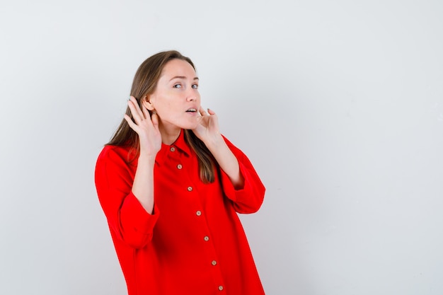 Jonge vrouw die handen achter de oren in rode blouse houdt en nieuwsgierig kijkt, vooraanzicht.