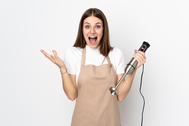 Jonge vrouw die handmixer gebruikt die op wit met verrassingsgelaatsuitdrukking wordt geïsoleerd