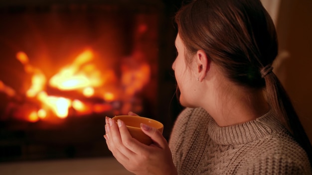 Jonge vrouw die hete thee drinkt en 's nachts in huis opwarmt bij brandend vuur in open haard