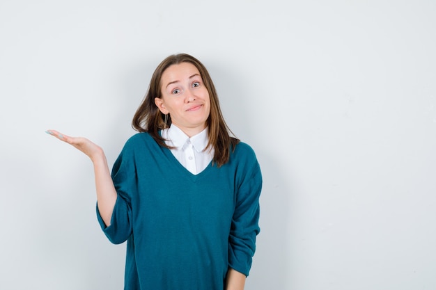 Jonge vrouw die hulpeloos gebaar toont met één hand in trui over wit overhemd en verward, vooraanzicht.