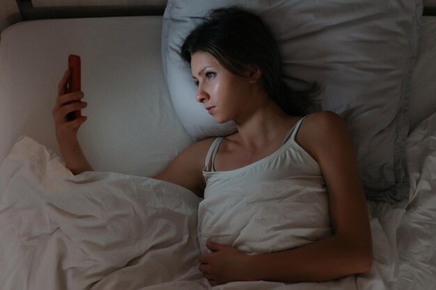 Jonge vrouw die in bed ligt en haar smartphone 's nachts gebruikt. Geen gezonde slaap. Afhankelijkheid van smartphones en sociale netwerken. Telefoon- en internetverslavingsconcept