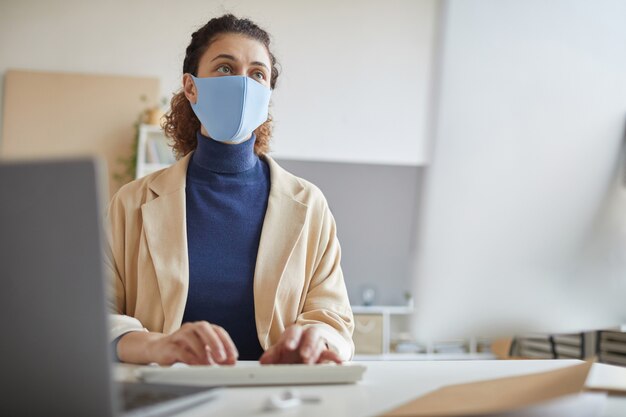 Jonge vrouw die in beschermend masker computermonitor bekijkt die zij online aan de lijst werkt tijdens pandemie