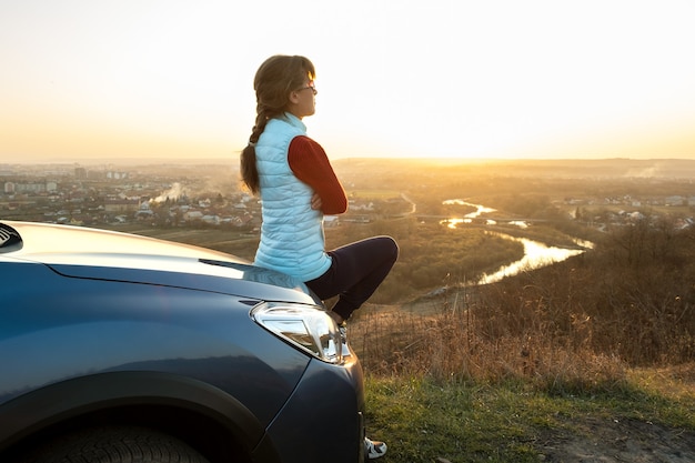 Jonge vrouw die in de buurt van haar auto staat en geniet van een warm uitzicht op de zonsondergang.