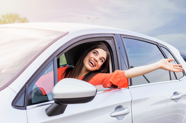 Jonge vrouw die in een auto zit met haar hand uit het raam Gelukkige vrouw die een auto rijdt en glimlacht Portret van gelukkige vrouwelijke chauffeur die een auto bestuurt met veiligheidsgordel Leuke jonge vrouw die gelukkig een auto rij