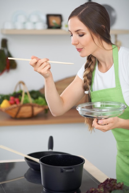 Jonge vrouw die in een keuken kookt Huisvrouw die soep proeft met een houten lepel