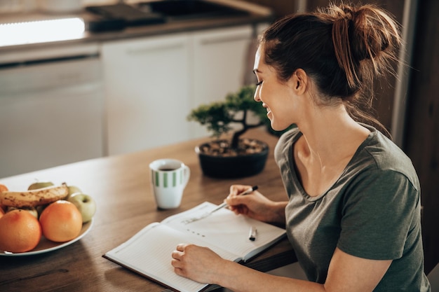 Foto jonge vrouw die in een notitieboekje schrijft en een takenlijst maakt terwijl ze thuis geniet van koffie in de ochtend.