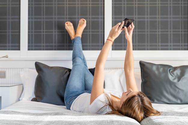 Jonge vrouw die in haar bed ligt dat haar mobiele telefoon met behulp van