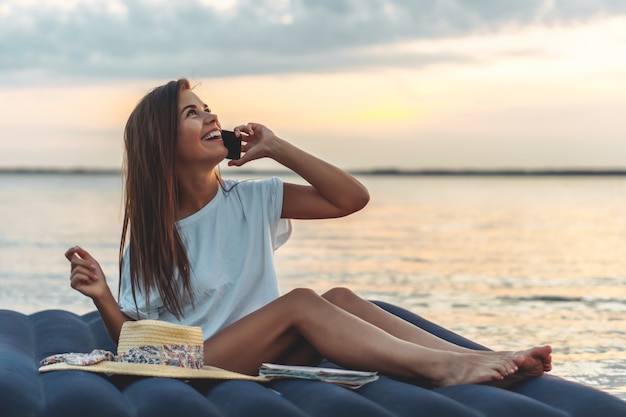 Jonge vrouw die in strohoed aan de smartphone op een strand spreekt.