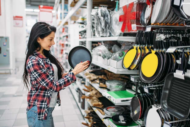 Jonge vrouw die koekenpan in supermarkt kiest. Vrouwelijke klant bij het winkelen in hypermarkt, producten voor de keuken