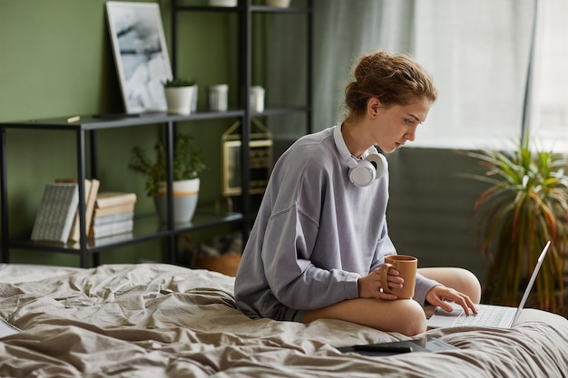 Jonge vrouw die koffie drinkt en 's ochtends thuis een laptop gebruikt terwijl ze op haar bed zit