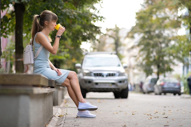 Jonge vrouw die koffie drinkt uit een papieren beker zittend op een stadsbank in het zomerpark