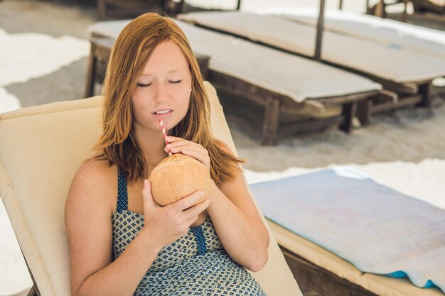 Jonge vrouw die kokosmelk drinkt op Chaise-longue op het strand.