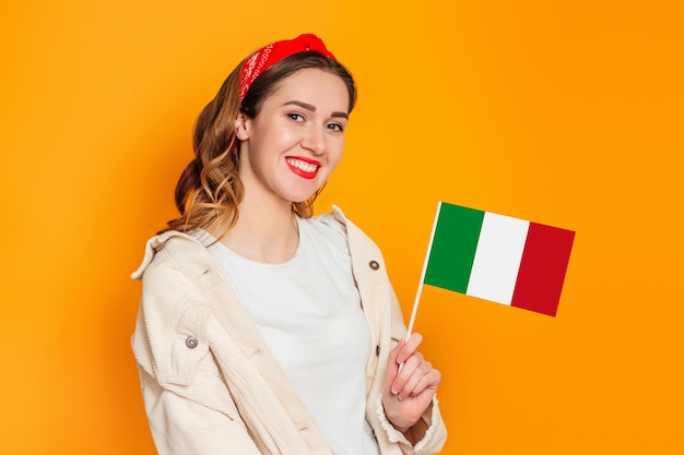 Jonge vrouw die lacht en de kleine vlag van Italië vasthoudt, geïsoleerd op een oranje achtergrond in de studio