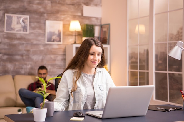 Jonge vrouw die lacht terwijl ze op laptop werkt en een kopje koffie vasthoudt. Vriendje ontspannen op de bank op de achtergrond.