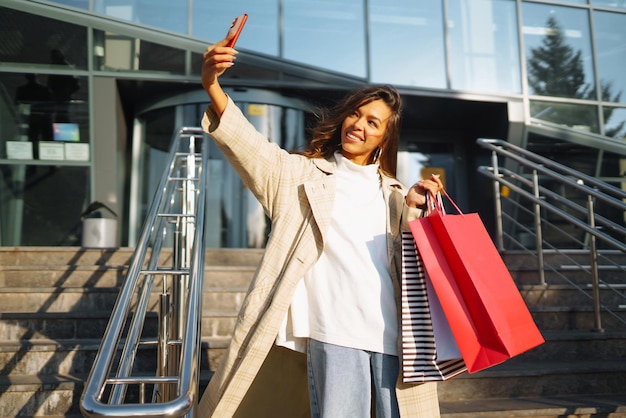 Jonge vrouw die langs de weg staat met boodschappentassen met mobiele telefoon Online winkelconcept