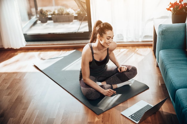 Jonge vrouw die laptop gebruikt terwijl ze 's ochtends thuis aan fitness doet.