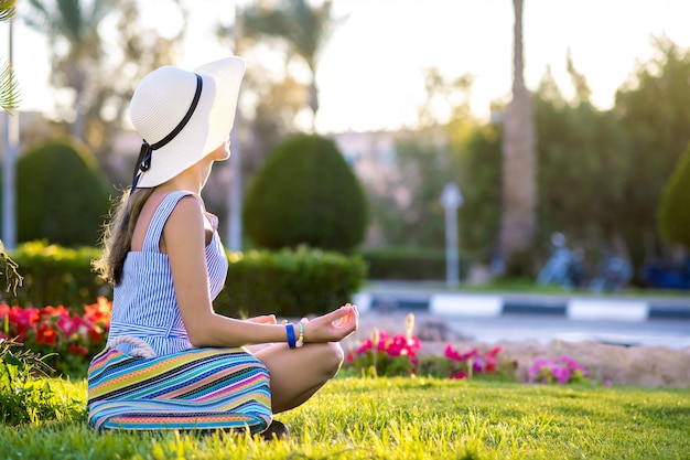 Jonge vrouw die lichtblauwe de zomerkleding en het gele strohoed ontspannen op groen grasgazon dragen in de zomerpark. Meisje in casual outfit rust buiten genieten van vrije tijd in warme ochtend.