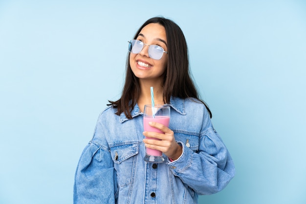 Jonge vrouw die met aardbeimilkshake over geïsoleerd blauw omhoog kijkt terwijl het glimlachen