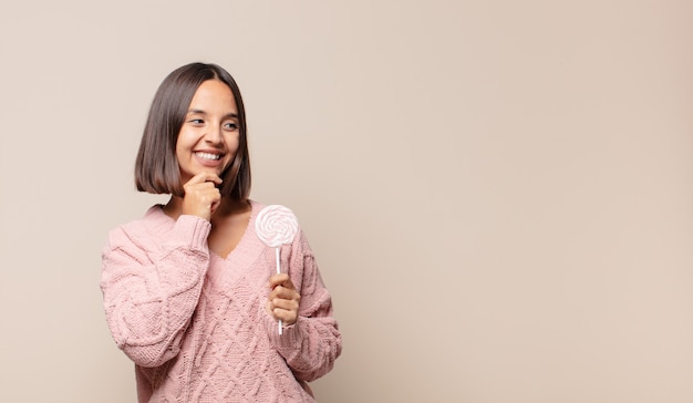 Jonge vrouw die met een gelukkige, zelfverzekerde uitdrukking glimlacht met hand op kin, zich afvraagt en naar de kant kijkt