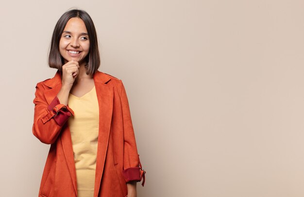 Jonge vrouw die met een gelukkige, zelfverzekerde uitdrukking glimlacht met hand op kin, zich afvraagt en naar de kant kijkt