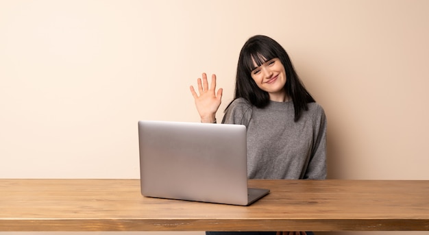 Jonge vrouw die met haar laptop die groeten met hand met gelukkige uitdrukking werkt
