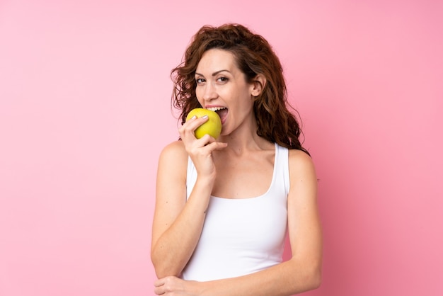 Jonge vrouw die met krullend haar een appel over geïsoleerd roze houdt