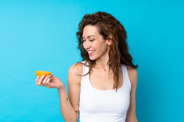 Jonge vrouw die met krullend haar een sinaasappel over geïsoleerd blauw houdt