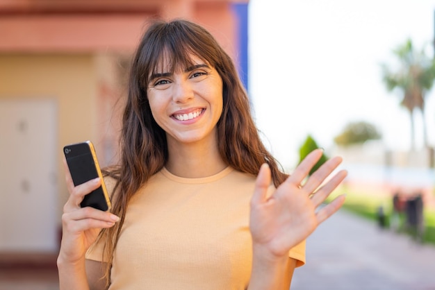 Jonge vrouw die mobiele telefoon gebruikt bij openlucht die met hand met gelukkige uitdrukking groeten