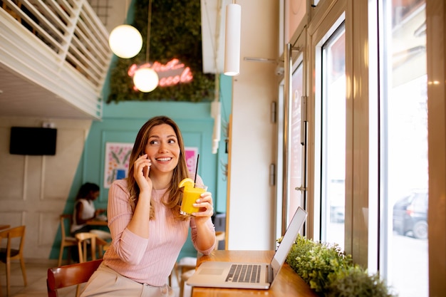 Jonge vrouw die mobiele telefoon gebruikt terwijl ze aan een laptop werkt en verse jus d'orange drinkt in het café