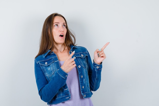 Jonge vrouw die naar de rechterbovenhoek wijst, omhoog kijkt in t-shirt, jas en zich afvraagt. vooraanzicht.