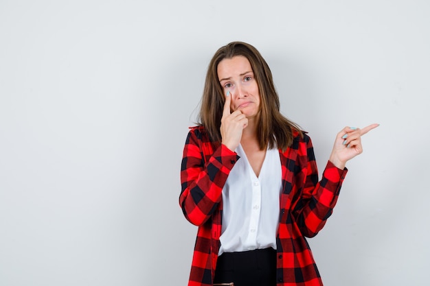 Jonge vrouw die naar rechts wijst en haar ooglid in vrijetijdskleding en er bedroefd uitziet, vooraanzicht.