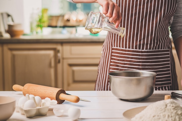 Jonge vrouw die olie in een kom met deeg giet, close-up. een vrouw in een gestreept schort kookt in de keuken