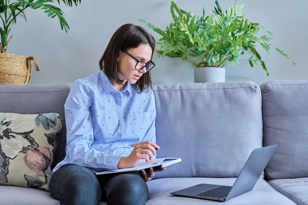 Jonge vrouw die online praat met behulp van laptop voor chatconferentie videogesprek notities maken terwijl ze thuis op de bank zit Vrijetijdswerk op afstand zakelijk onderwijs leren onderwijs technologie concept