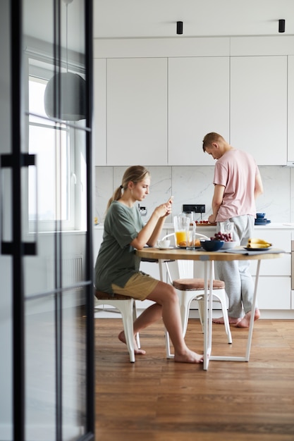 Jonge vrouw die ontbijt heeft en smartphone gebruikt terwijl haar echtgenoot het koken