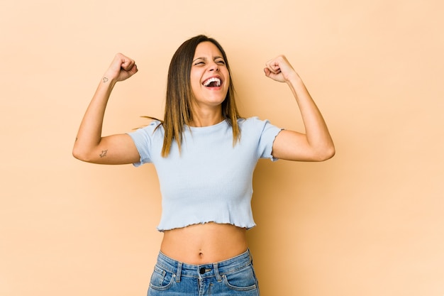 Jonge vrouw die op beige ruimte wordt geïsoleerd die vuist opheft na een overwinning, winnaarconcept.