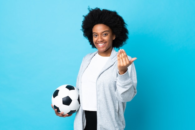 Jonge vrouw die op blauw met voetbalbal wordt geïsoleerd en komend gebaar doet