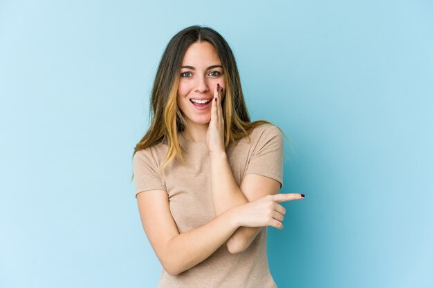 Jonge vrouw die op blauwe muur wordt geïsoleerd die een roddel zegt, naar kant wijst die iets meldt