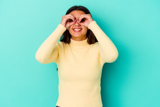 Jonge vrouw die op blauwe muur wordt geïsoleerd die ok teken over ogen toont