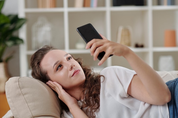 Jonge vrouw die op de bank ontspant en berichten leest op sociale media op het smartphonescherm