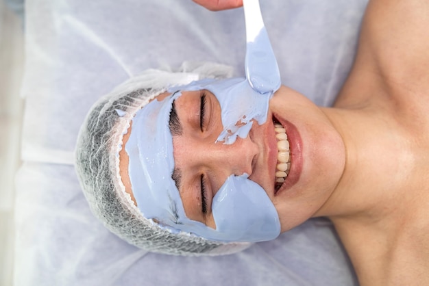 Jonge vrouw die op een massagetafel ligt en een blauw cosmetisch masker ontvangt in de schoonheidskliniek