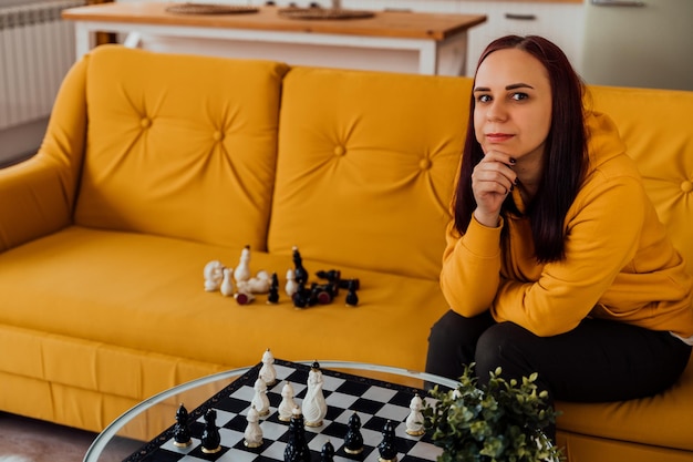 Jonge vrouw die op gele bank zit en schaak speelt in de kamer Vrouw speelt in logisch bordspel met zichzelf