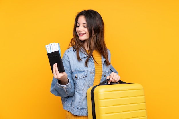 Jonge vrouw die op gele muur in vakantie met koffer en paspoort wordt geïsoleerd