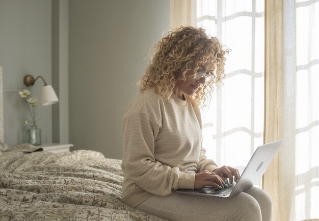 Foto jonge vrouw die op laptop werkt terwijl ze op bed in de slaapkamer zit. zakenvrouw die vanuit huis werkt. jonge vrouw in bril typen op laptop zittend op comfortabel bed in modern appartement