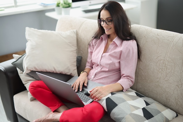 Jonge vrouw die op laptop werkt terwijl ze op de bank zit