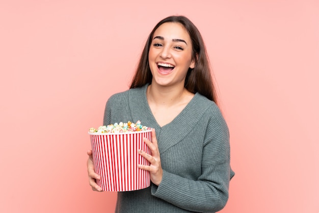 Jonge vrouw die op roze muur wordt geïsoleerd die een grote emmer popcorns houdt